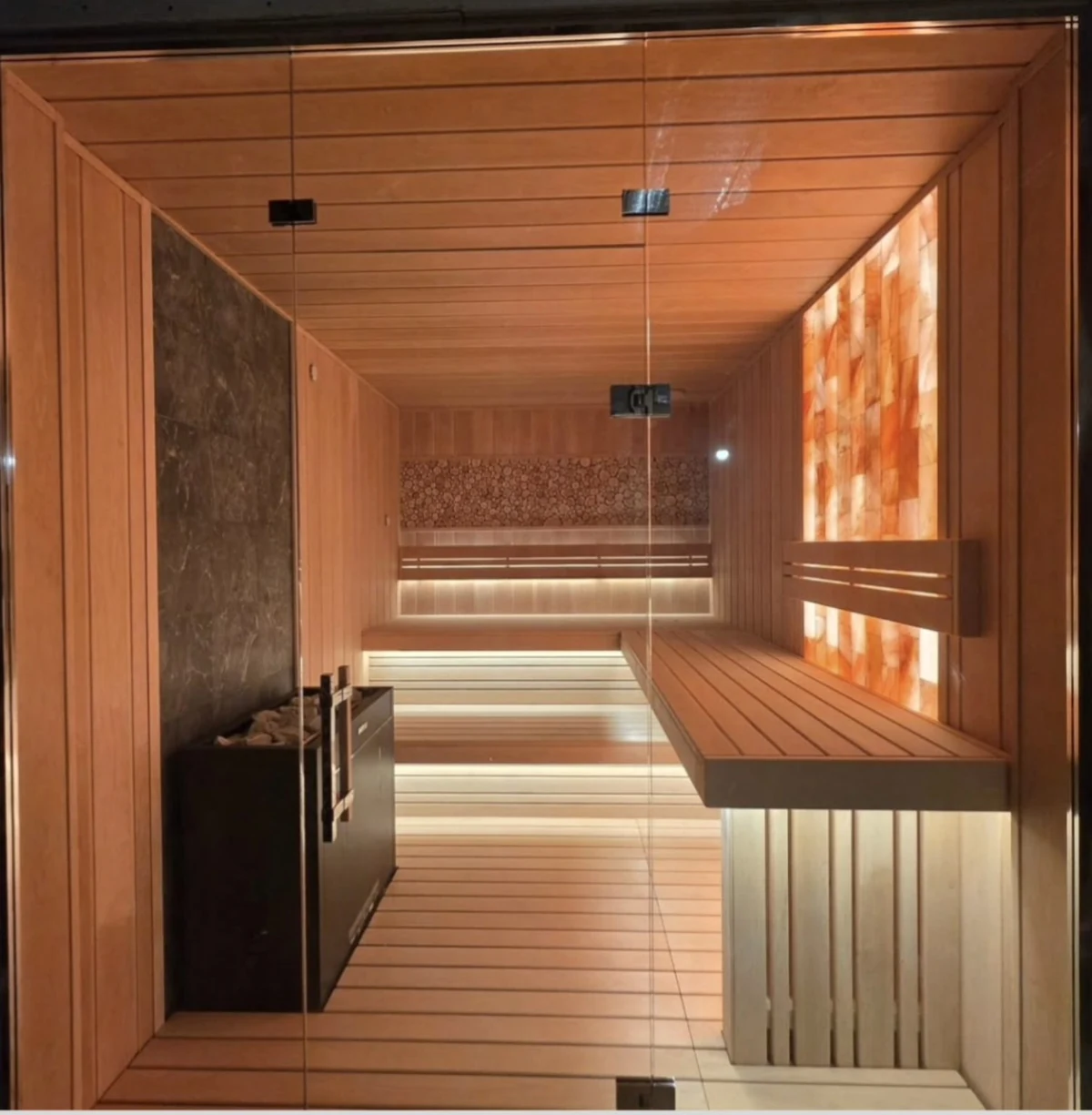 Glass-front sauna with L-shaped benches, an illuminated Himalayan salt wall and a black heater